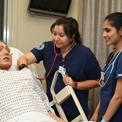 Two nursing students smile while checking on a simulation mannequin's breathing.